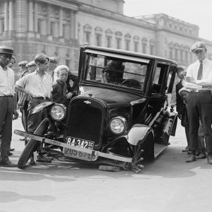 A Vintage Car Crash Attended By A Group Of Onlookers In A 1920s Urban Setting.