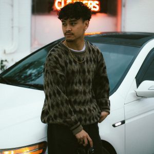 A Young Man Stands By A Car With A Camera Outside An Urban Building.