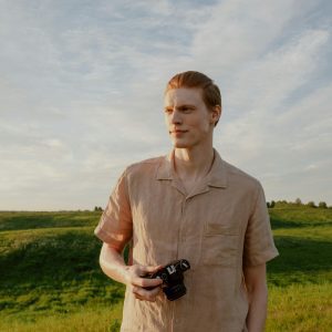 Redheaded Man Holding An Analog Camera In A Sunny Field With Blue Skies.