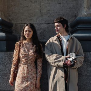 Two Stylish Individuals Pose With An Analog Camera By A Stone Wall In Daylight.