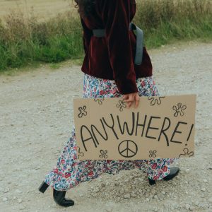 A Woman In A Vintage Outfit Holding A Sign Reading 'ANYWHERE!' On A Rural Road.