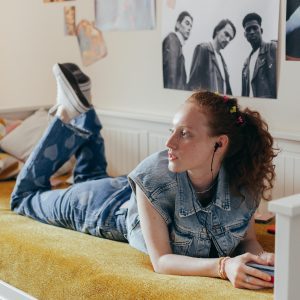 Red Haired Woman In Denim Vest Enjoying Music On Bed, Surrounded By Posters.