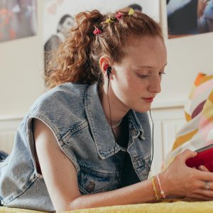 Teenage Girl In Denim Vest Listening To Music On A Retro Cassette Player While Lying On Bed.