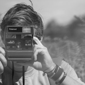 Black And White Photo Of A Man Holding A Vintage Polaroid Camera Outdoors.