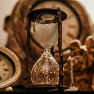 Close Up Of A Vintage Hourglass Surrounded By Antique Clocks In Warm Lighting.