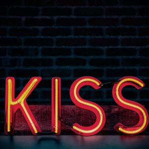 Bright Neon Lit 'Kiss' Sign With A Dark Brick Wall Background, Ideal For Modern Decor Themes.