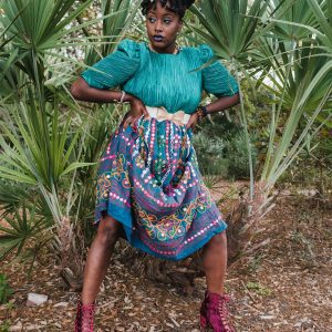 A Woman In A Colorful Dress With Afro Puffs Stands Confidently In A Tropical Garden.