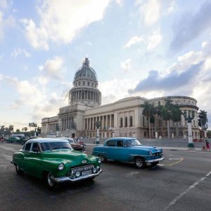 Classic Cars Drive Past El Capitolio In Havana, Showcasing Vintage Cuban Architecture And Urban Life.