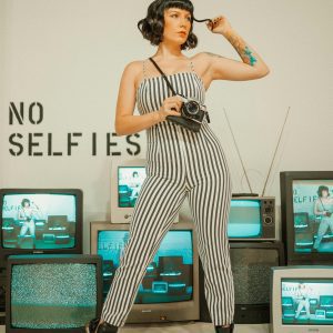 Fashionable Woman In Striped Jumpsuit Posing With Vintage Camera Amid Retro TVs. No Selfies Sign In Background.