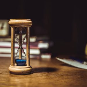 A Close Up Of A Wooden Hourglass With Blue Sand On A Wooden Desk, Symbolizing Time And Patience.