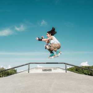 Woman Performs A High Jump On Roller Skates At An Outdoor Skate Park Under A Clear Blue Sky.