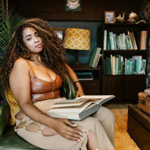 A Fashionable Woman Sitting With An Open Book In A Retro Inspired Room Featuring A Typewriter And Vintage Decor.