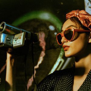 Woman In Polka Dot Attire Filming With A Vintage Camcorder, Exuding Retro Style.