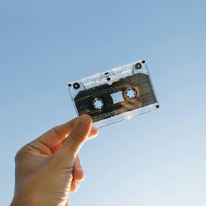 A Person Holds A Vintage Cassette Tape Against A Clear Blue Sky, Symbolizing Nostalgia And Technology.