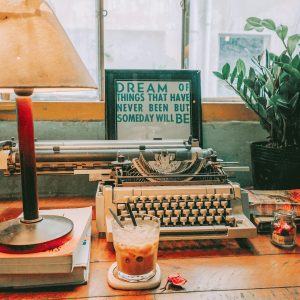 A Cozy Vintage Workspace Featuring A Typewriter, Lamp, And Iced Coffee.