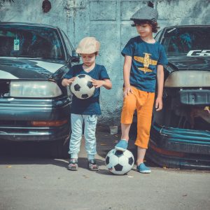 Two Children In Casual Attire Play Soccer Near Vintage Cars On A Sunny Day.