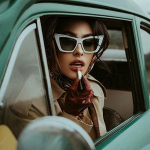 Stylish Woman Wearing Sunglasses And Applying Lipstick In A Classic Car, Embodying Timeless Fashion.