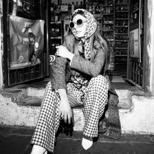 Caucasian Woman In Retro Fashion With Sunglasses Sitting At Store Entrance.