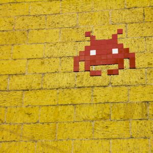 Street Art Featuring A Retro Pixelated Space Invader On A Vivid Yellow Brick Wall.