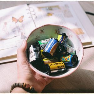 A Hand Holding A Bowl Filled With Various Vintage Film Rolls On A Sunlit Table.