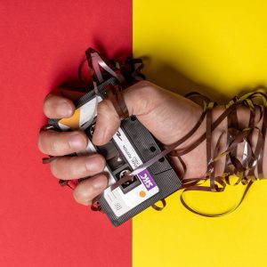 Hand Holding Tangled Cassette Tape On Vibrant Red And Yellow Background, Representing Nostalgia.