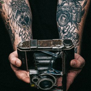 Close Up Of Hands Holding A Vintage Camera With Tattooed Arms.