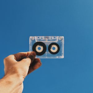 A Hand Holding A Transparent Vintage Cassette Tape Against A Clear Blue Sky, Capturing Nostalgia.
