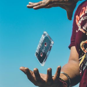 A Cassette Tape Floating Between Hands Against A Clear Blue Sky, Symbolizing Nostalgia And Retro Vibes.