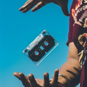 A Retro Cassette Tape Floating Between Hands Against A Clear Blue Sky, Evoking Nostalgia.