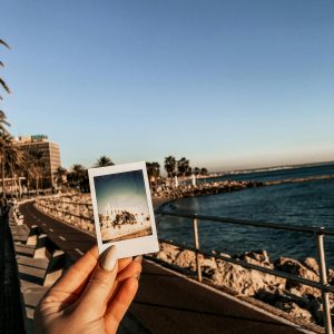 A Scenic Coastal Walkway With A Polaroid Picture In Hand Against A Sunset Backdrop.