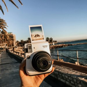 Close Up Of A Hand Holding An Instax Camera With Seaside View And Palm Trees.