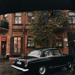 Classic Black Car Parked On A Rainy Street Amidst Urban Buildings.