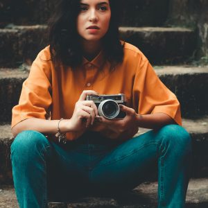 Stylish Woman Sitting On Stairs Holding A Vintage Camera, Showcasing Casual Wear And A Candid Pose.