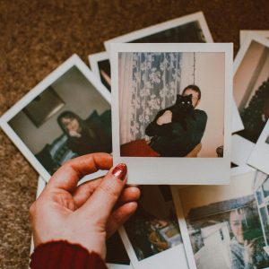 A Hand With Red Nails Holds A Vintage Polaroid Among Scattered Photos On Carpet.