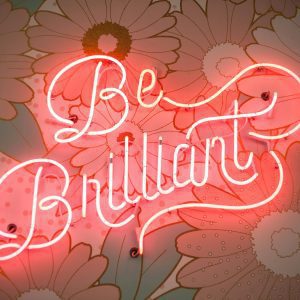 A Glowing Neon Sign Reading 'Be Brilliant' Against A Floral Backdrop Creates A Vibrant And Motivational Atmosphere.