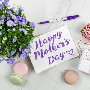 Flat Lay Of Mother's Day Card With Purple Flowers, Gift, Macarons, And A Pen Indoors.