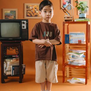 Asian Boy Standing In A Vintage Inspired Room With Orange Decor, TV, Books.