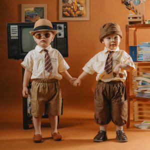 Two Boys In Elegant Vintage Attire Standing Indoors Holding Hands With An Orange Background.
