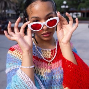 Stylish Young Woman Wearing Vibrant Sweater And Red Sunglasses Poses Confidently Outdoors.