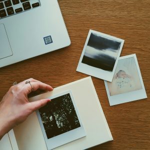 Hand Arranging Instant Photos Into An Album On Wooden Desk With Laptop.