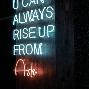 Neon Sign Stating 'U CAN ALWAYS RISE UP FROM ASHES' On A Dark Background, Glowing Brightly.