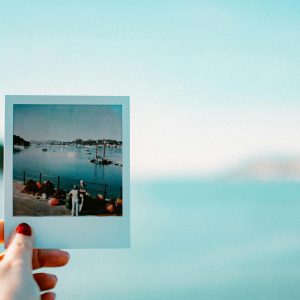 A Hand Holds A Polaroid Photo Of A Serene Waterfront View, With A Background Of Calm Waters And A Clear Sky.