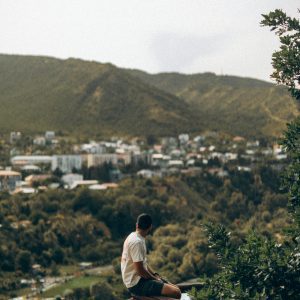 Man Sitting On Railing With Scenic View Of Tbilisi Hills And Cityscape.