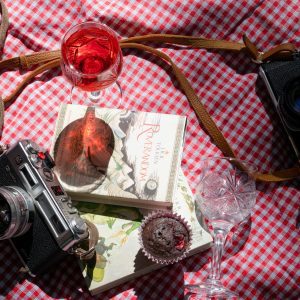 Charming Vintage Style Picnic Scene Featuring Cameras, Book, Wine, And Dessert On A Checkered Cloth.