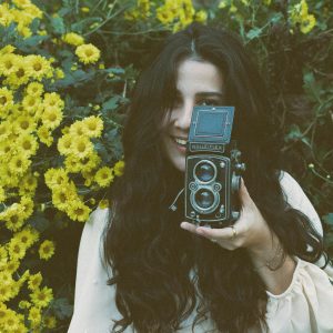 A Woman Holding A Rolleiflex Camera Amidst Vibrant Yellow Flowers In A Garden Setting.