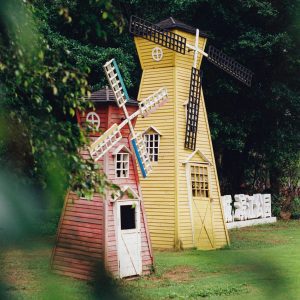 Two Wooden Windmills Stand In A Green Park Setting, Surrounded By Lush Trees.