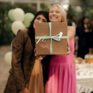 Two Women At A Festive Party Presenting A Gift Box With Balloons In The Background.