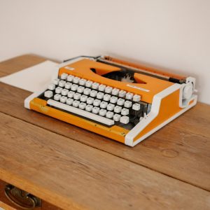 Retro Orange Typewriter Placed On A Rustic Wooden Desk In A Minimalist Setting.