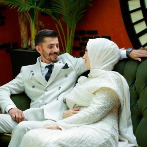 A Couple In Elegant Attire, Smiling And Seated On A Vintage Green Sofa Indoors With Vibrant Decor.