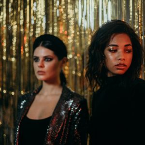 Two Women In Glamorous Outfits At A Party With Glittering Decorations, Exuding Fashion And Elegance.